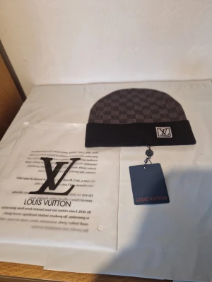 Louis Vuitton rutig mössa i svart - Snygg mössa från Louis Vuitton med klassiskt rutigt mönster i mörkgrått och svart. Mössan har en bred svart kant och LV-logga framtill. Perfekt accessoar för dig som gillar exklusiv streetstyle och vill sticka ut med designerplagg.