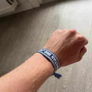 Blå Christian Dior armband med tofsar - Snyggt blått armband från Christian Dior med vit text och detaljer. Armbandet har ett randigt mönster och avslutas med flätade tofsar som ger en cool touch. Perfekt accessoar för dig som vill sticka ut med designerstil.