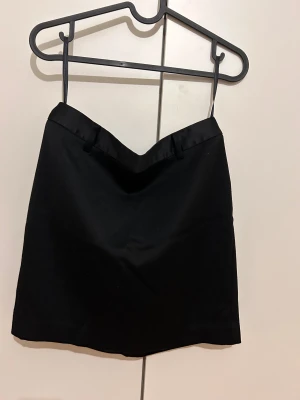 Svart kortkjol från Next, storlek 36 - Svart kortkjol från Next i storlek 36. Kjolen har en stilren design med slät yta och ett glansigt band upptill med hällor för bälte. Materialet känns mjukt och har en lätt struktur, perfekt för dig som gillar minimalistisk stil.