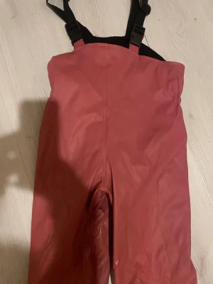 Röda täckbyxor från Exani med hängslen - Säljer ett par röda täckbyxor från Exani med justerbara svarta hängslen och reflexdetaljer vid bensluten. Byxorna är tillverkade i slitstark polyester och har hög midja för extra skydd mot kyla och väta. Perfekt för kalla dagar utomhus.