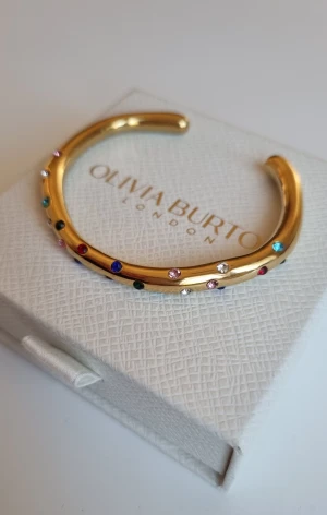 Guldigt armband med stenar - Snyggt guldigt armband, dekorerat med små färgglada stenar i blått, rött, grönt, rosa och vitt. Armbandet har en öppen design och en rundad form som gör det enkelt att ta på. Perfekt för dig som gillar glittriga detaljer och färg. Lådan ingår ej.