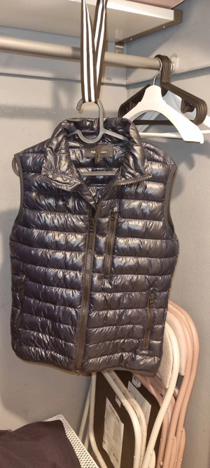 Mörkblå dunväst Peak Performance - Snygg mörkblå dunväst från Peak Performance med glansig finish och horisontella quiltade sömmar. Västen har hög krage, tre dragkedjeförsedda fickor och en diskret logga på ryggen. Perfekt för lager-på-lager och chill stil.