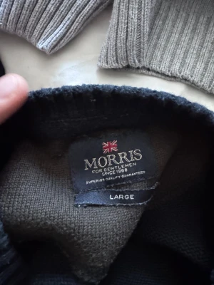 Mörkblå stickad tröja Morris L - Snygg mörkblå stickad tröja från Morris i storlek Large. Tröjan har broderad text och brittisk flagga på bröstet med 'Morris 1968'. Rund hals och långa ärmar med ribbade muddar. Perfekt för dig som gillar klassisk stil med en modern touch.