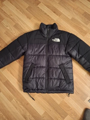 Svart pufferjacka The North Face - Säljer en svart pufferjacka från The North Face med hög krage och två snedställda fickor med dragkedja. Jackan har logga på bröstet och ryggen, quiltad design och justerbar nederkant med snörning. Perfekt för kalla dagar och har en clean look, är 175 och den passar väldigt bra I storlek.