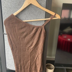 Brun ribbad oneshoulder topp - Säljer en brun ribbad topp med oneshoulder-design och smalt axelband. Toppen är tight och har en snygg asymmetrisk form som ger en cool vibe. Materialet är mjukt och stretchigt, perfekt för att styla med jeans eller kjol. Passar dig som gillar trendiga och enkla plagg.