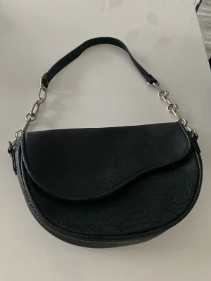 Svart axelväska med kedjedetaljer - Snygg svart axelväska med rundad form och strukturmönster som ger en cool look. Väskan har ett lock med magnetknäppning och en axelrem med silverfärgade kedjor. Perfekt storlek för det viktigaste och passar till många outfits.Kom priv för fler bilder plick laggar