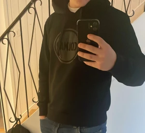 Svart hoodie från Colmar med logga - Svart hoodie från Colmar med stor logga i svart på bröstet. Tröjan har huva med snörning och ribbade muddar vid ärmslut och nederkant. Tillverkad i mjuk bomull och polyesterblandning, perfekt för chill dagar. Snygg och enkel att matcha med jeans eller joggers. Tröjan är lite liten i storleken och passar S bättre