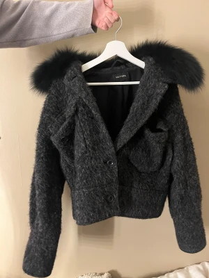 Svart teddyjacka med päls från Meotine - Säljer en svart teddyjacka från Meotine med fluffig päls på kragen. Jackan har en croppad passform, stora knappar framtill och en cool, lite oversized look. Materialet är mjukt och ulligt, perfekt för höst och vinter. Snygg detalj med ficka på bröstet.