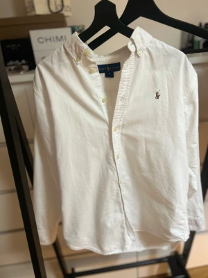 Vit skjorta från Ralph Lauren - Vit klassisk skjorta från Ralph Lauren med button-down krage och broderad logga på bröstet. Skjortan har långa ärmar och knappar framtill. Tillverkad i mjuk bomull som känns skön mot huden. Perfekt för dig som gillar stilrena och tidlösa plagg.