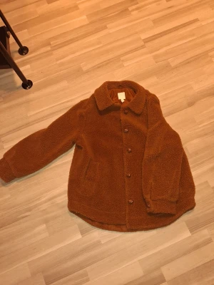 Brun teddyjacka från H&M  - Säljer en brun teddyjacka från H&M i storlek 42. Jackan har mjukt och fluffigt material, klassisk krage och knäppning med bruna knappar. Fodret är slätt och matchar jackans färg. Använd en gång sen har den bara legat i garderoben. 
