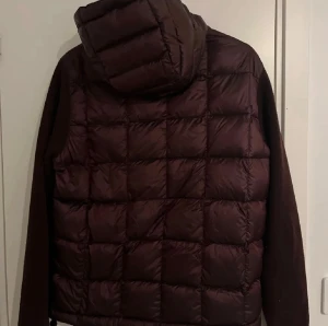 Vinröd pufferjacka med huva - Säljer en vinröd pufferjacka med quiltad framsida och rygg, samt stickade ärmar. Jackan har dragkedja framtill, två fickor med dragkedja och en stor huva. 