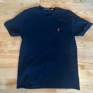 Mörk blå t-shirt från Polo Ralph Lauren - Svart t-shirt från Polo Ralph Lauren med klassisk röd broderad logga på bröstet. Modellen är kortärmad och har rund hals. Tillverkad i mjuk bomull som känns skön mot huden. Perfekt för dig som gillar stilrena och ikoniska plagg. Använd 1 gång