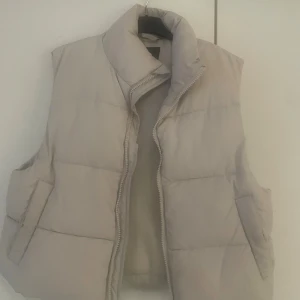 Beige dunväst - Snygg beige dunväst med quiltad design och hög krage. Västen har två stora fickor på sidorna och stängs med dragkedja framtill. Perfekt att slänga över en hoodie för extra värme. Enkel och stilren look som funkar till många outfits.