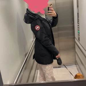 Svart dunjacka från Canada Goose - Svart dunjacka från Canada Goose  klassisk logga på ärmen. Jackan har dragkedja framtill, två fickor och ribbade muddar vid ärmslut. Perfekt för kalla dagar och har en stilren, lång modell som håller dig varm hela vintern. Tyvärr ingår inte pälsen då den är borttappad