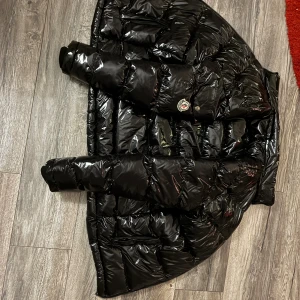 Svart pufferjacka från Moncler - Säljer en svart, glansig pufferjacka från Moncler med klassisk logga på bröstet. Jackan har lång ärm, quiltad design och dragkedja framtill. Perfekt för kalla dagar och har en riktigt snygg oversized look. Materialet är syntet och dun, vilket gör den både varm och lätt.