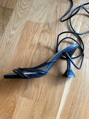 Svarta strappy pumps från Lovit - Snygga svarta pumps från Lovit med tunna remmar och fyrkantig tå. Skon har en unik, skulptural klack och långa snören som kan lindas runt vristen. Tillverkade i konstläder med en stilren och trendig design som passar perfekt till många outfits.