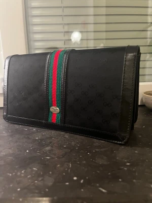 vintage Svart Gucci  kuvertväska med röd/grön detalj - ( den har en skråma på framsidan )Snygg svart kuvertväska från Gucci med diskret GG-mönster och klassiska röd-gröna ränder i mitten. Väskan är rektangulär och har detaljer i läder samt en liten metallbricka framtill. Perfekt för dig som gillar stilrena accessoarer med lyxig känsla.