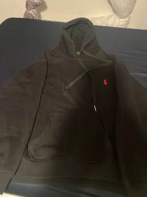 Svart hoodie från Ralph Lauren - passar i size M oxå. Snygg svart hoodie från Ralph Lauren med klassisk röd broderad logga på bröstet. Hoodien har huva med dragsko och en stor magficka framtill. Perfekt för dig som gillar stilrena och tidlösa plagg.