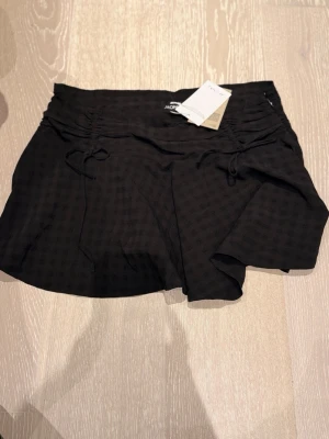 Kjol från Monki - Supersnygg svart kjol från Monki  med diskret rutmönster och dragsko i midjan. Kjoldesign framtill och shorts under för extra komfort. Perfekt för dig som vill ha en trendig och bekväm look. Materialet känns lätt och följsamt.