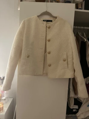 Krämvit bouclé kavaj från Zara - Stilren krämvit kavaj från Zara i bouclétyg med rund halsringning och fyra dekorativa guldknappar framtill. Kortare modell med raka ärmar och två fickor fram. Perfekt för en chic och trendig look.