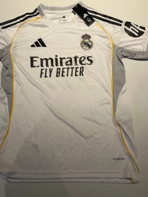 Real Madrid matchtröja Adidas S - Vit Real Madrid matchtröja från Adidas med svarta och gula detaljer. Tröjan har klubbmärke, sponsortryck och Aeroready-material som andas. Slim fit-modell med kort ärm och sportig design, perfekt för fotbollsträning eller match.