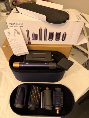 Dyson Airwrap Complete Long hårstyler - Dyson Airwrap Complete Long i mörklila och koppar med flera stylingtillbehör. Inkluderar olika borstar och cylindrar för att skapa lockar, volym och släta frisyrer. Kommer i en elegant förvaringsväska. Perfekt för kreativ hårstyling med avancerad teknik.