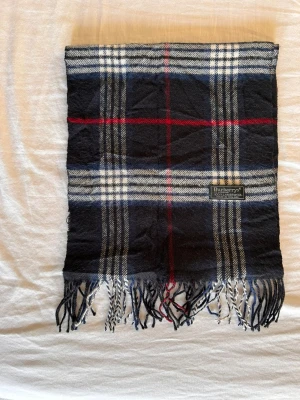 Rutig halsduk från Burberry - Snygg halsduk från Burberry i klassiskt rutigt mönster med mörkblå, vita och röda ränder. Halsduken har fransar i ändarna och är tillverkad i mjukt material som känns lyxigt och stilrent. Perfekt accessoar för att lyfta din outfit.