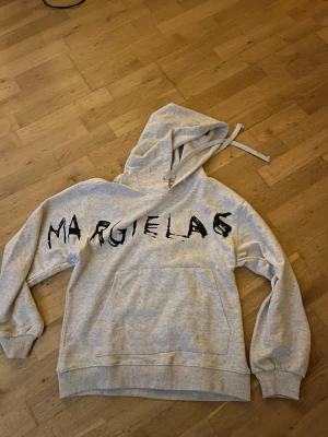 Grå hoodie från Margiela - Snygg grå hoodie från Margiela med stor svart text på bröstet. Hoodien har huva med dragsko och en stor magficka framtill. Tillverkad i mjukt bomullsmaterial och har ribbade muddar vid ärmar och nederkant. Perfekt för en avslappnad och trendig look. Den är lite losse fit. 