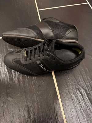 Svarta BOSS sneakers - Snygga svarta sneakers från BOSS med stilren design. Skorna har en kombination av läder och textil, svarta skosnören och diskret logga på sidan. Perfekta för dig som gillar en clean och sportig look. Strlk 42, passar också er med 43
