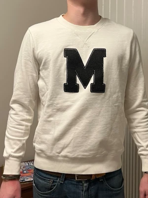 Vit sweatshirt Från Morris - Vit sweatshirt med stort svart M-tryck på bröstet. Tröjan har rund halsringning, ribbade muddar och är tillverkad i mjukt material. Perfekt för en avslappnad och stilren look. Har lite små ljus gråa pricka på baksida armar, men inget man lägger märke till utan noggrann titt. 