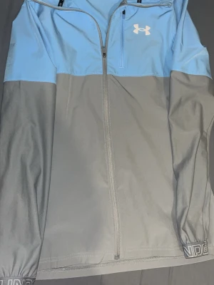 Blågrå vindjacka från Under Armour - Snygg vindjacka från Under Armour i ljusblått och grått. Jackan har hel dragkedja, bröstficka med dragkedja och elastiska muddar med logga på ärmsluten. Perfekt för blåsiga dagar och sportiga outfits.