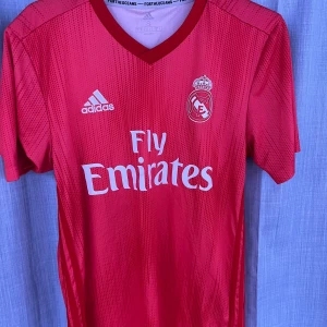 Röd Real Madrid matchtröja Adidas - Röd Real Madrid matchtröja från Adidas med vitt tryck och klubbmärke på bröstet. Tröjan har korta ärmar, V-ringning och är tillverkad i ett lätt, ventilerande funktionsmaterial. Perfekt för fotbollsträning eller sportiga outfits.