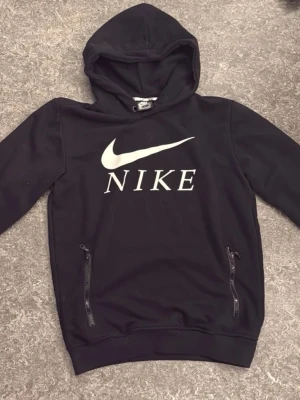 Svart Nike Hoddie med fickor - Snygg svart hoodie från Nike med stor vit logga och text på bröstet. Hoodien har huva och två praktiska fickor med dragkedja framtill. Tillverkad i mjukt material som passar perfekt till en avslappnad streetwear-look. Storlek M