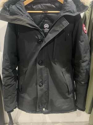 Svart dunjacka från Canada Goose (äkta) - Säljer en svart dunjacka från Canada Goose med det klassiska röda och vita Arctic Program-märket på ärmen. Jackan har en stilren design och är perfekt för kalla vinterdagar. Fylld med dun för maximal värme och komfort. Äkta!