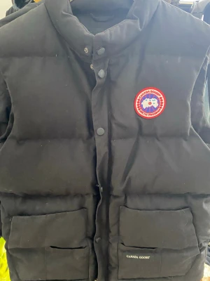 Svart dunväst från Canada Goose - Säljer en svart dunväst från Canada Goose med klassisk logotyp på bröstet och två stora fickor framtill. Västen har hög krage, tryckknappar och dragkedja. Tillverkad i slitstarkt material och fylld med dun för extra värme. Perfekt för kalla dagar.