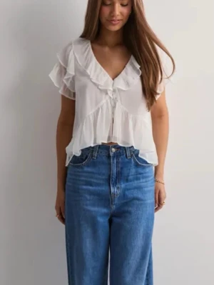 Svart volangblus med v-ringning - Söt svart blus med volanger och djup v-ringning. Blusen har korta ärmar och knappar framtill. Köpt från Nelly (hittade inga bilder på den svarta på hemsidan men det är exakt samma blus) OBS! Första knappen går inte att knäppa då den är sönder men vill man stänga går det med en säkerhetsnål. 