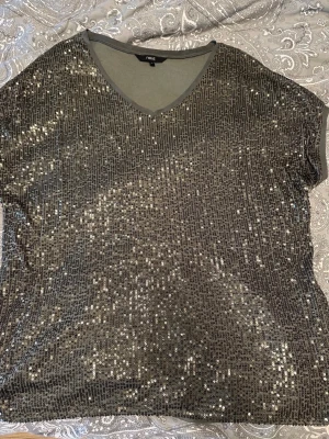 Next Glittrig topp, storlek 38/40 - Glittrig topp från Next med V-ringning och korta ärmar. Perfekt för festliga tillfällen. Den är ny, endast provat. Den passar storlek 38/40   Rök och djurfritt hem
