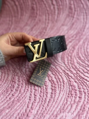 Svart  skärp med guldspänne - Snyggt svart skärp från  med präglat monogrammönster och stort  i guld. Bältet är brett och tillverkat i skinn, perfekt för att lyfta vilken outfit som helst. Ikonisk accessoar för dig som gillar lyxiga detaljer.