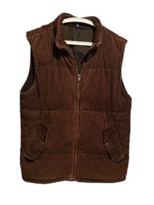 Brun manchester dunväst med dragkedja - Snygg brun dunväst i manchestertyg med dragkedja framtill och två fickor med lock. Västen har en klassisk krage och quiltad design som ger en cool och avslappnad look. Perfekt för lager-på-lager-stil under kyliga dagar.