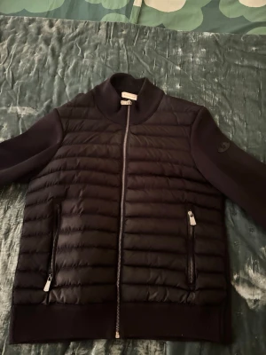 Svart cardigan från Moncler - Snygg svart cardigan från Moncler med quiltad front och två dragkedjeförsedda fickor. Västen har ribbade detaljer vid krage och nederkant samt en diskret Moncler-logga på ärmen. Perfekt för lager-på-lager och streetwear-vibe.