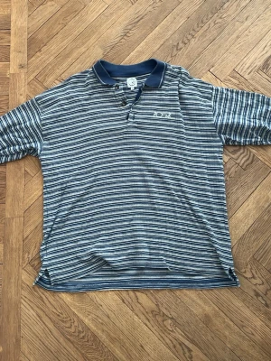 Randig polotopp från Polar Skate Co - Snygg randig polotopp från Polar Skate Co i blått och vitt. Klassisk pikékrage med två knappar och broderad logga på bröstet. Tillverkad i mjuk bomull, perfekt för en avslappnad stil.