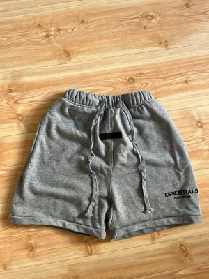 Grå shorts från Essentials Fear of God - Gråa shorts från Essentials Fear of God med elastisk midja och snörning framtill. Mjukt material och logga tryckt på vänster ben. Perfekta för en avslappnad och trendig stil.