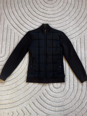 Svart pufferjacka med dragkedja - Stilren svart pufferjacka med quiltat mönster och hög krage. Jackan har hel dragkedja framtill, två sidofickor med tryckknappar och släta ärmar. Perfekt för kalla dagar och enkel att matcha med olika outfits. Stolek 2 (S)
