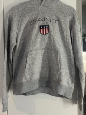 Grå hoodie från GANT med logga - Snygg grå hoodie från GANT med broderad logga och sköld på bröstet. Klassisk känguruficka framtill och huva. Perfekt för en avslappnad och sportig stil. Mjuk och bekväm bomullsblandning.
