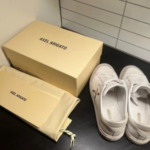 Vita sneakers från Axel Arigato - Stilrena vita sneakers från Axel Arigato med lågt skaft. Skorna har en minimalistisk design med subtil logga på sidan och ovandel i läder och mocka. Klassiska vita snören och diskret beige sula ger en clean look som passar till det mesta. Skorna passar även väldigt fint till sommaren och våren! Skriv om ni har några frågor🫡😄