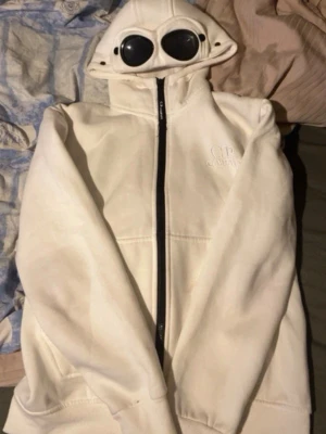 Vit hoodie med goggles från C.P. Company - Säljer en unik vit hoodie från C.P. Company med ikoniska goggles i huvan och dragkedja framtill. Hoodien har en clean look med diskret logga på bröstet och är tillverkad i mjukt material. Perfekt för dig som vill sticka ut med streetwear-vibe.
