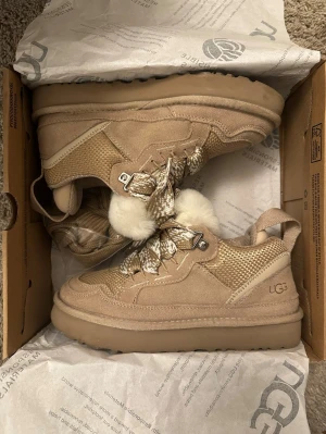 UGG Lowmel sneakers  - Snygga beige sneakers från UGG med chunky sula, stickade och mockadetaljer samt coola snören med fuskpäls. Perfekta för dig som vill ha en unik och trendig look. UGG-logga på sidan och bakre hälflik för enkel påtagning. Använd endast 1 gång. Färgen är Sand!!