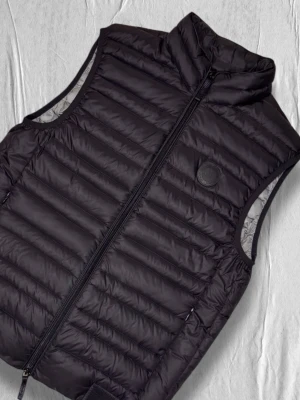 Svart dunväst från Armani Exchange - Stilren svart dunväst från Armani Exchange med quiltad design och hög krage. Västen har dragkedja framtill, diskret logotyp på bröstet och ljusgrå insida. Perfekt för lager-på-lager och ger en modern look.