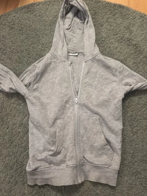 Grå zip hoodie från Kappahl - En enkel och stilren grå hoodie från Kappahl med dragkedja framtill och två fickor. Hoodien har en klassisk huva och är gjord i mjukt sweatshirtmaterial, perfekt för chill dagar. Passar dig som gillar basic och bekvämt.