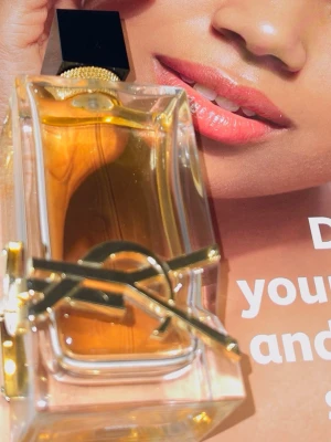 Yves Saint Laurent Libre Eau de Parfum - Säljer en lyxig parfym från Yves Saint Laurent, Libre Eau de Parfum. Flaskan är rektangulär i genomskinligt glas med guldiga detaljer och en svart kork. Innehållet är bärnstensfärgat och flaskan pryds av YSL:s ikoniska logga i metall.